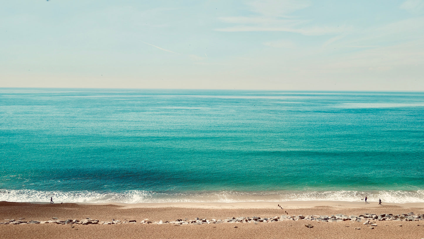 A photographic view of Barton Beach from atop the cliffs. A calm sea and lightly clouded sky with dog walkers enjoying the beach. 